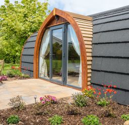 An outdoor patio area with wooden chairs and table next to a pod with arched glass doors and a garden bed with flowers at Chapel Pod in Callington