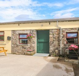 An outdoor area with a stone and yellow wall building with three windows and a green door a wooden picnic table and flower pots at The Old Cows House in Helston