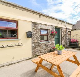 An outdoor patio area with a wooden picnic table and a potted plant a stone and plaster wall with windows a green door and flower baskets at The Old Cows House in Helston