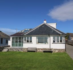 A single-story house with white walls and a dark roof a wooden bench and table on the lawn at Angorfa - Trearddur Bay