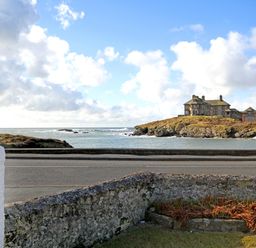 A coastal view with a stone wall in the foreground a road and a stone building on a rocky hill at Angorfa - Trearddur Bay in Trearddur Bay