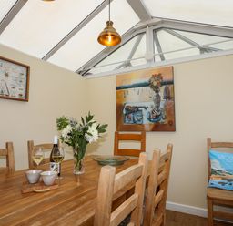 A dining area with a wooden table and chairs a vase of flowers and framed pictures on beige walls at Arkoo in Rhosneigr
