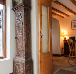 A wooden carved grandfather clock next to a glass door leading to a room with a wooden chair a desk and a lamp at Barras Cottage in Brynsiencyn