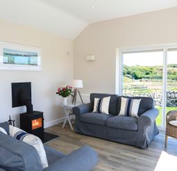 A living room with two gray sofas, a wicker chair, a small wood-burning stove, and large windows with a view at Bwthyn Glanrhyd in Llanfair yn Neubwll