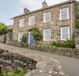 A stone house with a blue door and white framed windows by a paved road and stone walls at Bron Castell in Harlech