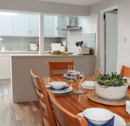 A dining table set with plates glasses and a plant with a kitchen area in the background at Brook Cottage in Criccieth