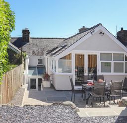 A patio area with a table and six chairs outside a house with white walls and a sloped roof at Brook Cottage in Criccieth