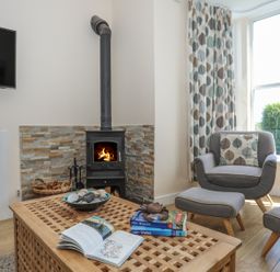 A living room with a wood stove fireplace a wooden coffee table with books and shells grey armchairs with leaf patterned cushions and curtains at Bryn Dale in Trearddur Bay