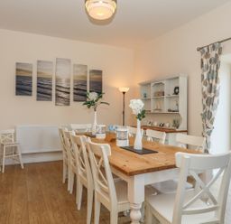 A dining room with a wooden table and white chairs next to a window with patterned curtains at Bryn Dale in Trearddur Bay
