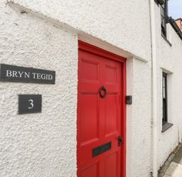 A white textured exterior wall with a red door and house sign reading Bryn Tegid number 3 in Beaumaris