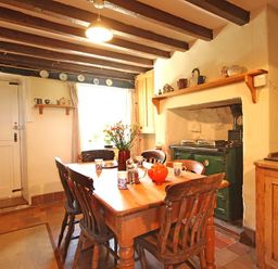 A kitchen with wooden dining table and chairs a green stove and shelves with crockery at Buck Cottage in Red Wharf Bay