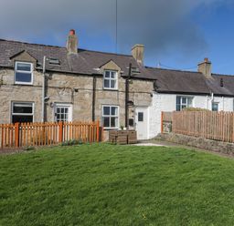 A row of connected stone and white-painted houses with chimneys and a grassy yard enclosed by wooden fences at Cae Person in Llangoed