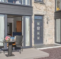 A patio area with two black chairs and a table with flowers outside a stone building with a door labeled Cambrian House in Criccieth