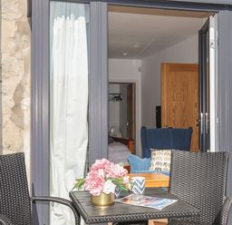 An outdoor seating area with two woven chairs and a table with flowers and mugs in front of open glass doors showing a room with a blue armchair and bed at Cambrian House in Criccieth