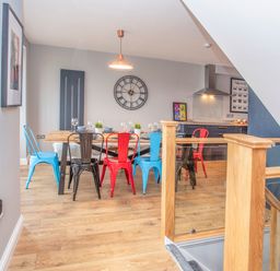 A dining area with a wooden table and multicolored metal chairs next to a kitchen and wooden staircase at Cambrian House in Criccieth