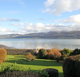 A garden with bushes and grass overlooking a body of water and hills in the background at Camlan in Beaumaris