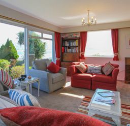 A living room with sofas and armchairs by large windows with a view of the sea and garden at Camlan in Beaumaris