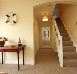 A hallway with a wooden table holding flowers and a decorative boat under a life ring on the wall and stairs to the right at Camlan in Beaumaris