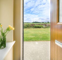 An open wooden door showing a concrete path and grass lawn with flowers in a glass vase on a white windowsill at Coed Bolyn Lodge in Bethel