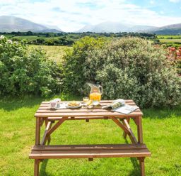 A wooden picnic table with mugs plates a jug of juice and a magazine in a garden with bushes and mountains in the background at Coed Bolyn Lodge in Bethel