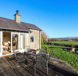 An outdoor patio with a metal table and four chairs next to a house with glass doors opening to a living room and green fields with mountains in the background at Cosy Barn in Llangaffo
