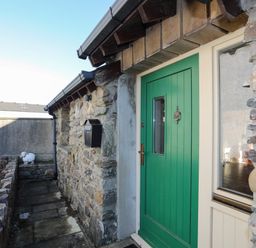 A stone exterior wall with a green door and a black mailbox beside a small stone path at Cosy Barn in Llangaffo
