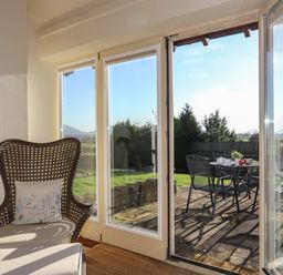 A wicker chair with a cushion next to glass doors opening to a patio with a table and metal chairs at Cosy Barn in Llangaffo