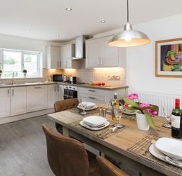 A kitchen with white cabinets and gray countertops and a wooden dining table with chairs set for meals and decorated with a plant at Cuckooland in Borth-Y-Gest