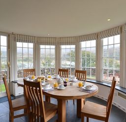 A dining area with a round wooden table and six chairs set for breakfast in a room with large windows at Dinas Noddfa in Nant Gwynant
