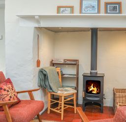 A sitting area with a wood-burning stove a chair with a blanket and shelves with magazines at Ffrwd Cottage in Bodorgan