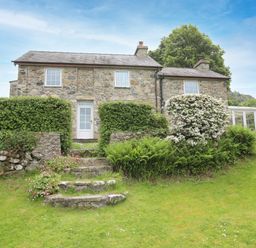 A stone cottage with a white door and windows surrounded by green bushes and a grassy lawn at Fron Fawr in Nefyn