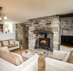 A living room with stone walls a fireplace with a wood stove wicker sofas a television and a wooden shelf at Fron Fawr in Nefyn