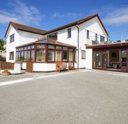 The exterior of a two story white house with a brown roof a sunroom with wooden framed windows a wooden bench and a patio area at The Gables in Trearddur Bay