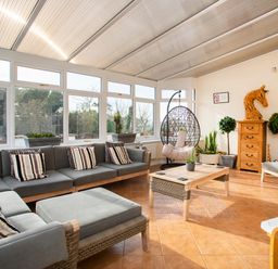 A sunroom with a sectional sofa, coffee table, hanging chair, wooden chest, and potted plants at The Gables in Trearddur Bay