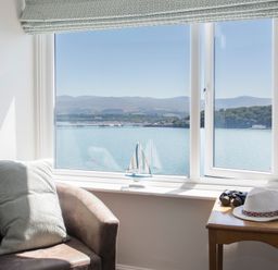A room with a chair, pillow, small wooden table with a hat and binoculars by a window overlooking water and hills at Glan Dwr Bach in Glyn Garth