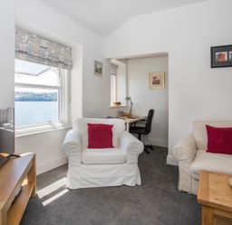 A living room with white sofas and red cushions a wooden table with fruit a TV on a wooden stand and a desk with a chair near windows at Glan Dwr Bach in Glyn Garth