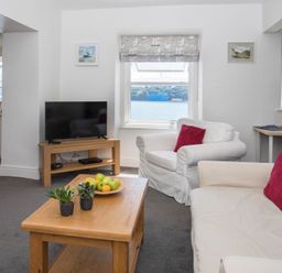 A living room with white armchairs and sofa with red cushions a wooden coffee table with plants and fruit a TV on a wooden stand and a dining area visible through an open doorway at Glan Dwr Bach in Glyn Garth