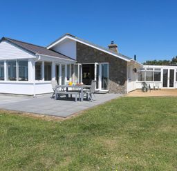 A white and stone house with a patio table and chairs on a paved area and plastic chairs on a wooden deck in a grassy yard at Glanrhyd Uchaf in Llanfair yn Neubwll