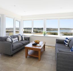 A living room with two grey sofas and a wooden coffee table by large windows overlooking a river at Glanrhyd Uchaf in Llanfair yn Neubwll