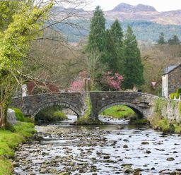 A stone bridge over a shallow river with trees and stone buildings on either side at Gwyndy Bach in Beddgelert