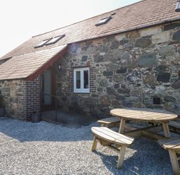 An outdoor area with a stone house wall a wooden picnic table and benches at Hafod in Pontllyfni