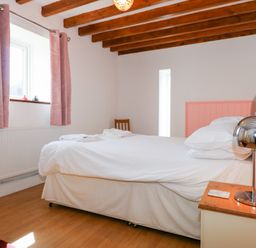 A bedroom with a bed with white linens a wooden nightstand with a lamp pink curtains a wooden chair and wooden ceiling beams at Hafod in Pontllyfni