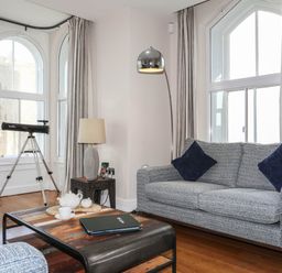 A sitting area with a gray sofa, a wooden coffee table with a teapot set, a telescope near large arched windows, and a floor lamp at Harlech Apartments - Branwen in Harlech
