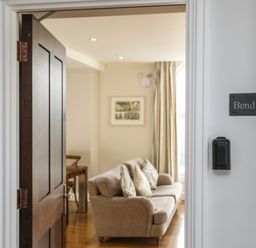 An open door revealing a living room with a beige sofa and cushions next to a window with curtains at Harlech Apartments - Bendigeidfran in Harlech