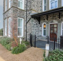 The exterior entrance of a stone building with a wooden door and iron fence at Harlech Apartments - Gwern in Harlech