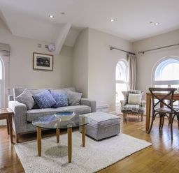 A living area with a grey sofa, patterned cushions, a glass coffee table, an armchair, a wooden dining table and chairs at Harlech Apartments - Matholwch in Harlech