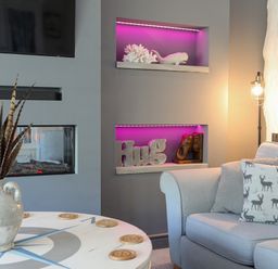 A living room with a grey sofa with deer printed cushions a table with a vase and feathers and a wall with two shelves lit in purple at Hen Gilfach in Llanfairyngnhornwy