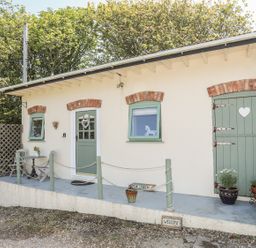 The exterior of a small building with green doors and windows and a small porch with chairs and potted plants at Hen Gilfach Stable in Llanfairyngnhornwy