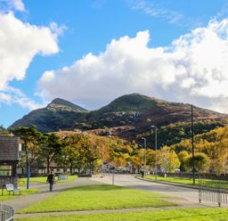 A park area with grass and benches along a road with mountains in the background at Hirfron in Llanberis