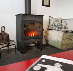 A room with a woodburning stove a metal kettle on a small table and a cushioned bench with fur throw and ship patterned pillows at Big House in Llandecwyn